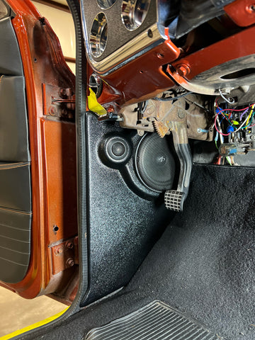 A black speaker enclosure installed in the kick panel area of a vehicle, with visible wiring and part of the car's interior.